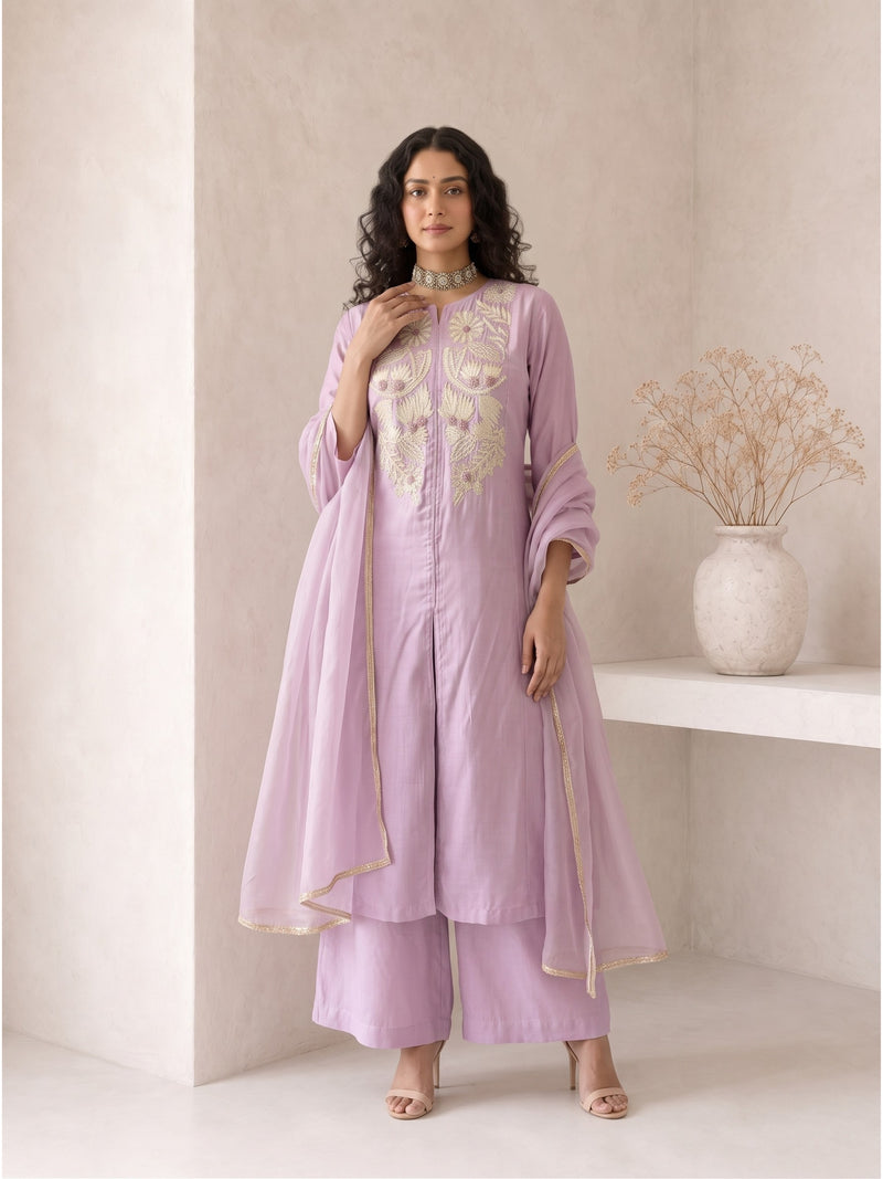 A woman models a lavender purple traditional Indian outfit consisting of a kurta with gold embroidery, matching palazzo pants, and a sheer dupatta, set against a minimalist beige wall with a shelf and dried flowers.