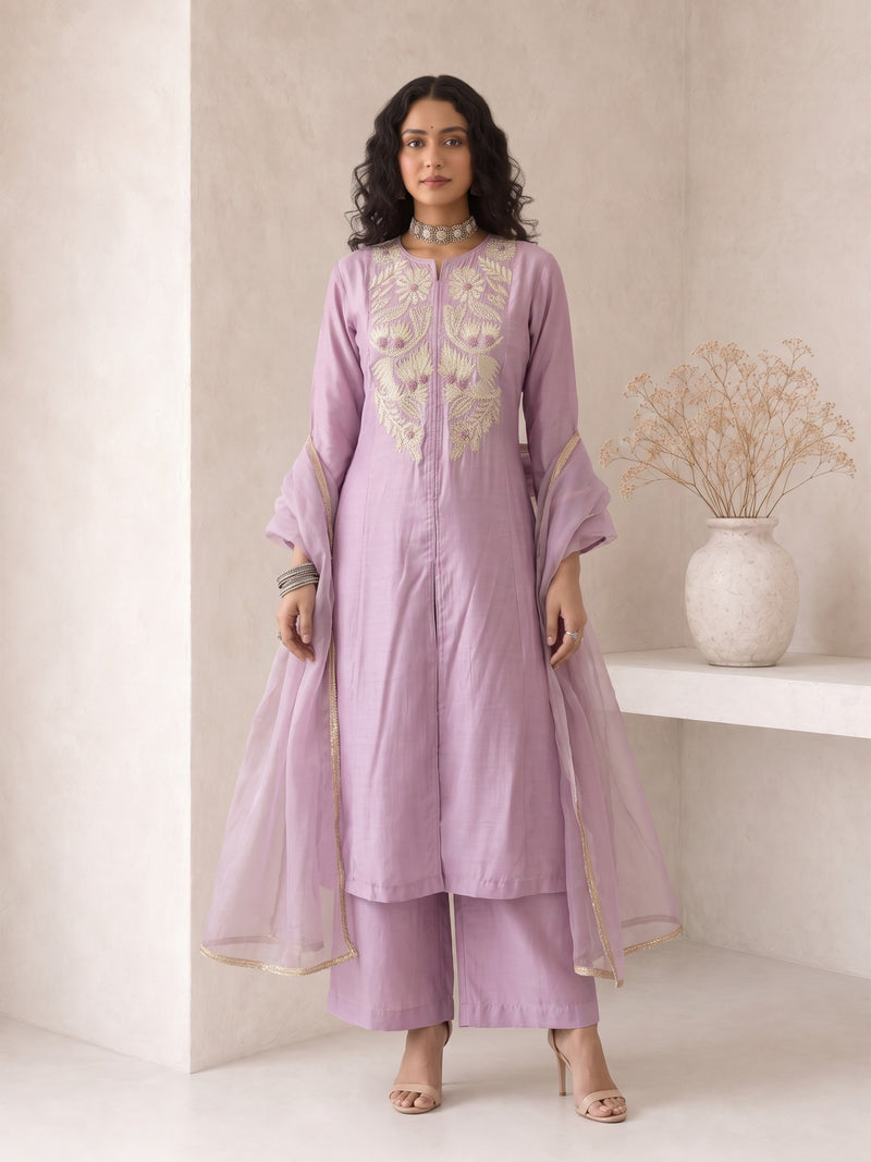 A woman models a lavender purple traditional Indian outfit consisting of a long kurta with gold embroidery, matching trousers, and a sheer matching dupatta, set against a minimalist beige wall with a decorative vase.