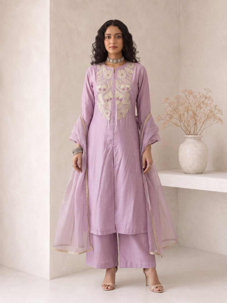 A woman models a lavender purple traditional Indian outfit consisting of a long kurta with gold embroidery, matching trousers, and a sheer matching dupatta, set against a minimalist beige wall with a decorative vase.