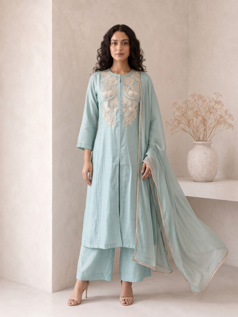 A woman models a light blue-green traditional Indian outfit consisting of a long kurta with gold embroidery, matching trousers, and a sheer dupatta, set against a minimalist beige wall with a decorative vase.