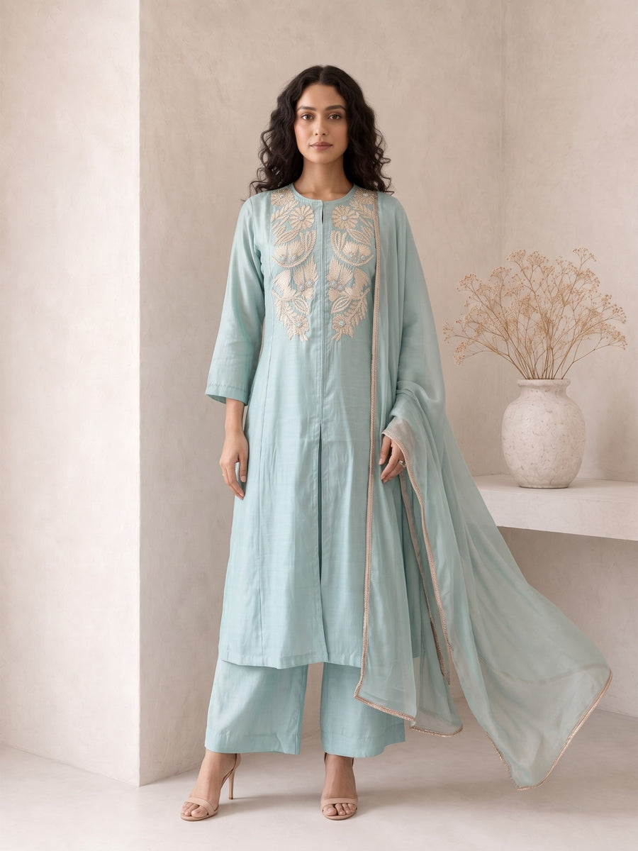 A woman models a light blue-green traditional Indian outfit consisting of a long kurta with gold embroidery, matching trousers, and a sheer dupatta, set against a minimalist beige wall with a decorative vase.