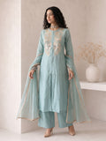 A woman models a light blue-green traditional outfit with beige embroidery on the chest, matching wide-leg pants, and a sheer dupatta with beige border trim, set against a minimalist beige wall with a decorative vase.