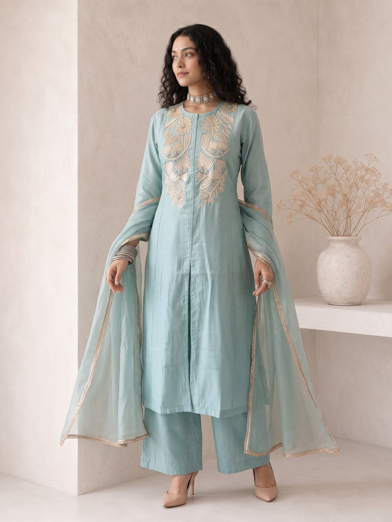 A woman models a light blue-green traditional outfit with beige embroidery on the chest, matching wide-leg pants, and a sheer dupatta with beige border trim, set against a minimalist beige wall with a decorative vase.