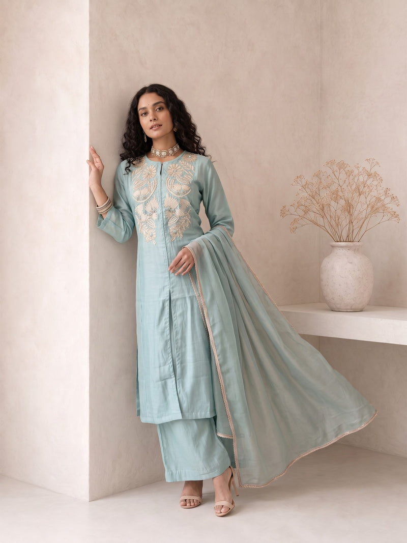 A woman models a light blue-green traditional Indian salwar kameez suit with beige embroidery on the chest, paired with matching trousers and a sheer dupatta, set against a minimalist beige wall background.